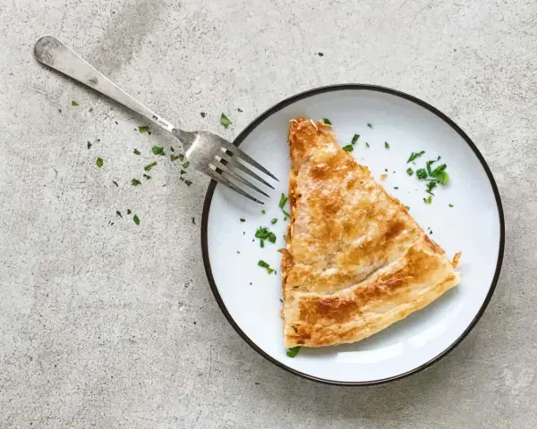 Una porción dorada de empanada de atún, lista para ser disfrutada con un tenedor plateado y espolvoreada con perejil fresco.