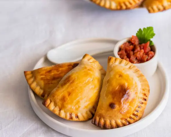 Tres empanadillas rellenas doradas y crujientes, servidas con una pequeña taza de salsa de tomate y una hoja de perejil.