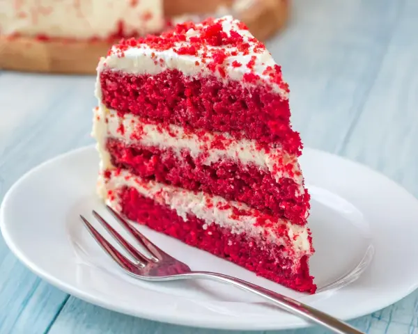 Rebanada de torta red velvet con glaseado blanco y migas rojas, servida en plato blanco con tenedor.