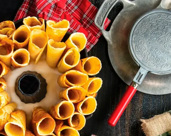 Deliciosos postres noruegos en forma de conos dorados, listos para rellenar. Una vieja gofrera de hierro reposa cerca.