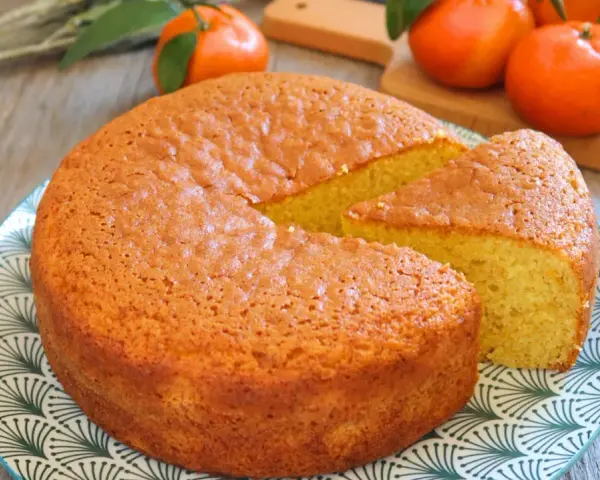 Bizcocho de mandarina y yogur, esponjoso y dorado, con una porción cortada. Mandarinas frescas al fondo.