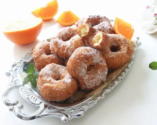 Rosquillas de naranja recién hechas, espolvoreadas con azúcar, junto a gajos de naranja fresca en una bandeja plateada.