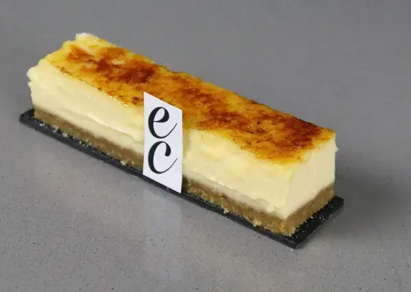 Un trozo de cheesecake creme brulee con base crujiente y cubierta caramelizada, adornado con un distintivo.