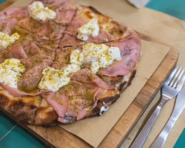 Pizza con mortadela y burrata cremosa, espolvoreada con hierbas verdes y servida en tabla de madera.