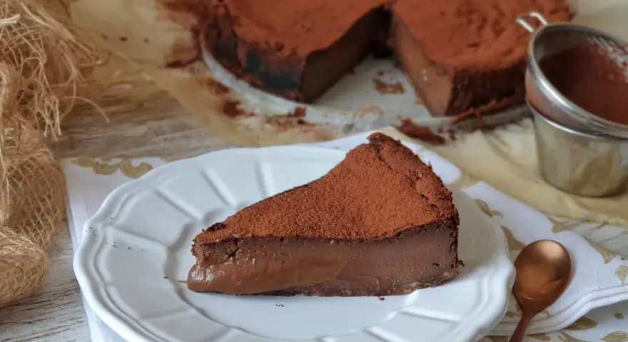 Una rebanada de torta de chocolate, cubierta de cacao en polvo, reposa en un plato blanco. El postre se ve cremoso y delicioso.