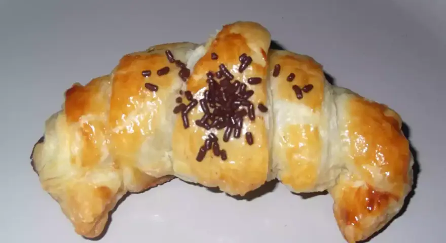 Croissant de chocolate recién horneado, dorado y brillante, espolvoreado con virutas de chocolate.