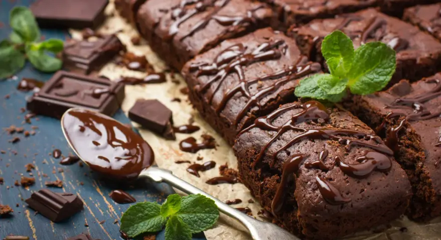 Deliciosos postres de chocolate, brownies cubiertos con glaseado de chocolate y decorados con menta fresca. Trozos de chocolate y una cuchara con chocolate derretido completan la escena.