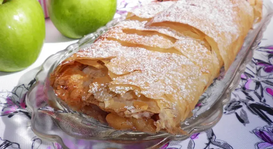 Delicioso strudel de manzana recién horneado, espolvoreado con azúcar glas y acompañado de manzanas verdes frescas y canela.