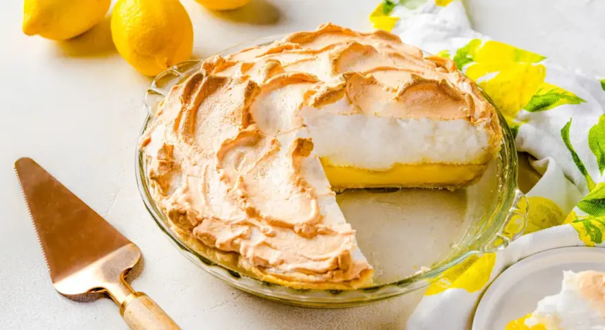 Tarta de limón con merengue dorado y esponjoso, lista para servir. Dos limones frescos y un paño con estampado de limones completan la escena.