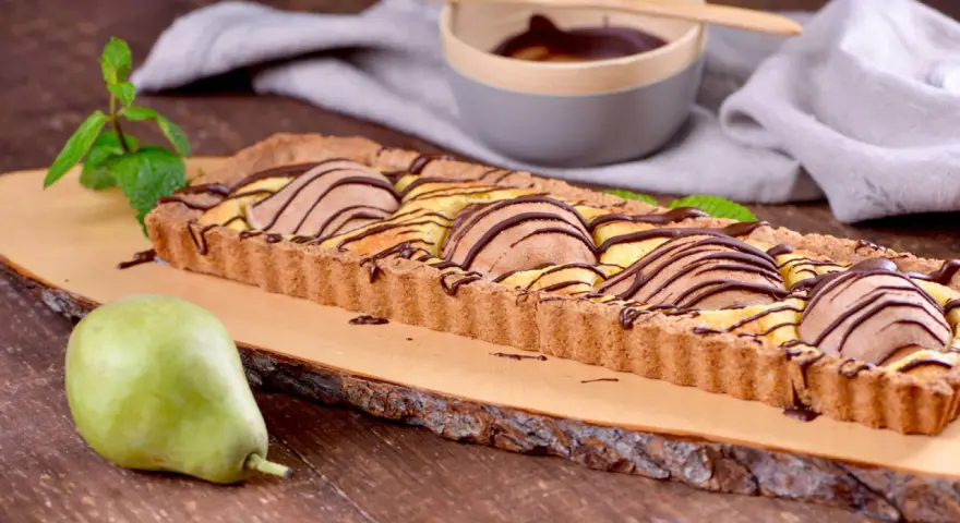 Tarta rectangular con peras y chocolate, decorada con menta. Una pera fresca reposa a un lado.
