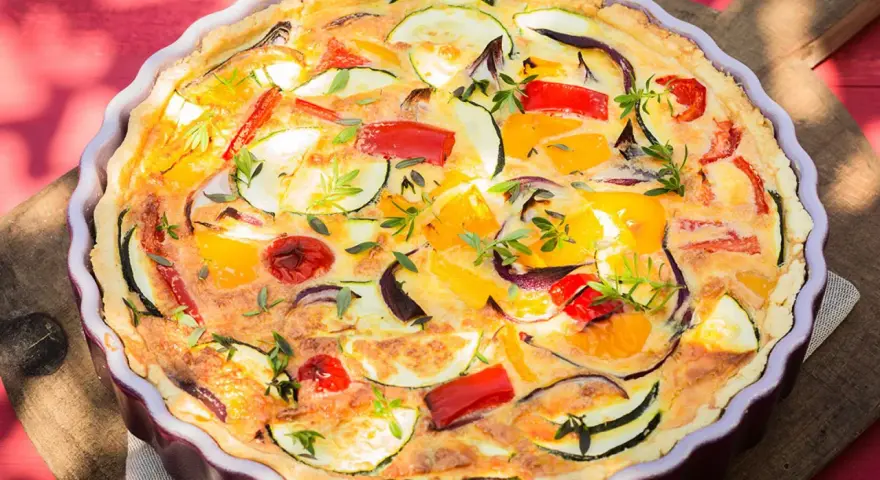 Quiche de verduras con hojaldre, repleta de pimientos, calabacín y cebolla morada, adornada con hierbas frescas.
