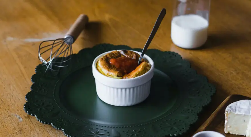 Un esponjoso soufflé con salsa de tomate, listo para disfrutar. Al lado, un batidor y un vaso de leche, ingredientes clave para crear que es un soufflé.