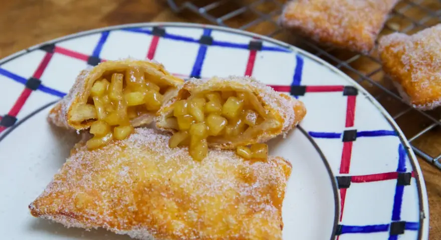 Empanada de manzana recién hecha, dorada y espolvoreada con azúcar. Una está partida, mostrando el relleno jugoso de manzana.