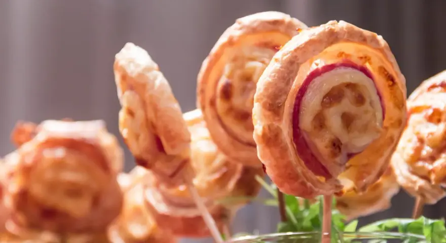 Deliciosas piruletas de hojaldre rellenas de queso y jamón, presentadas en un vaso con hojas verdes.