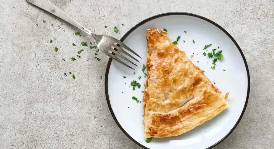 Una porción dorada de empanada de atún, lista para ser disfrutada con un tenedor plateado y espolvoreada con perejil fresco.