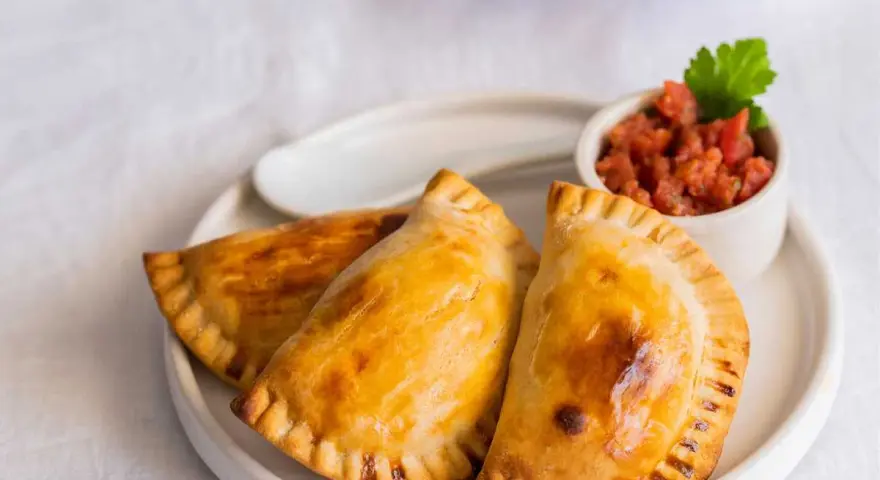 Tres empanadillas rellenas doradas y crujientes, servidas con una pequeña taza de salsa de tomate y una hoja de perejil.