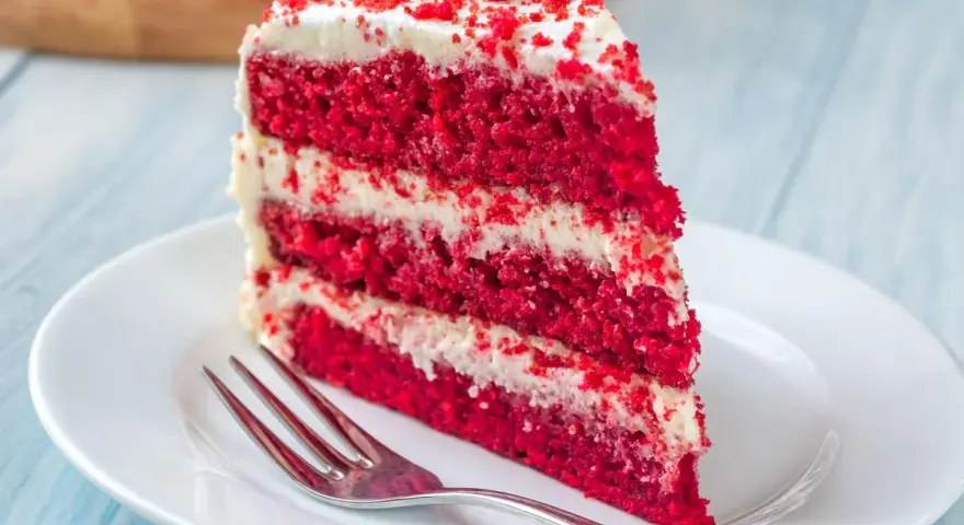 Rebanada de torta red velvet con glaseado blanco y migas rojas, servida en plato blanco con tenedor.