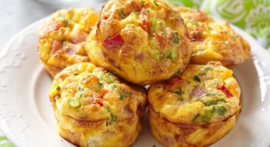 Montón de muffins saludables con huevo, pimiento rojo, cebolleta y jamón, espolvoreados con perejil.