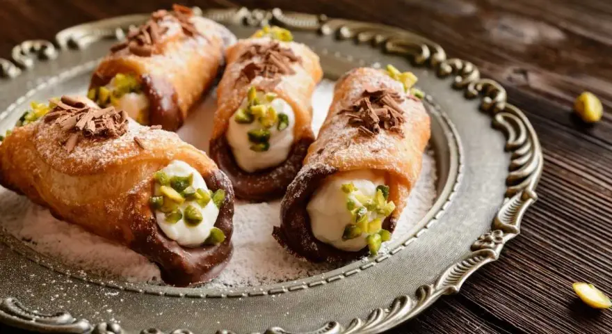 Cuatro cannoli siciliano, crujientes y rellenos de crema dulce, espolvoreados con pistachos y virutas de chocolate.