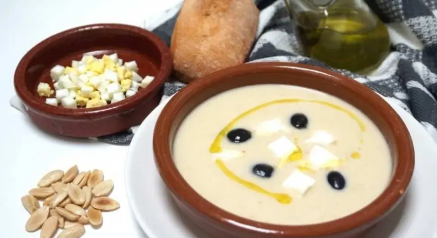 Un cuenco de mazamorra cordobesa con aceitunas, huevo duro y un chorrito de aceite. Acompaña un panecillo y almendras.