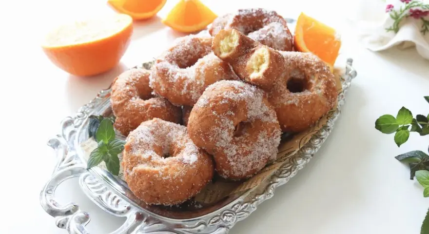 Rosquillas de naranja recién hechas, espolvoreadas con azúcar, junto a gajos de naranja fresca en una bandeja plateada.