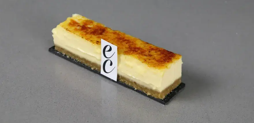 Un trozo de cheesecake creme brulee con base crujiente y cubierta caramelizada, adornado con un distintivo.
