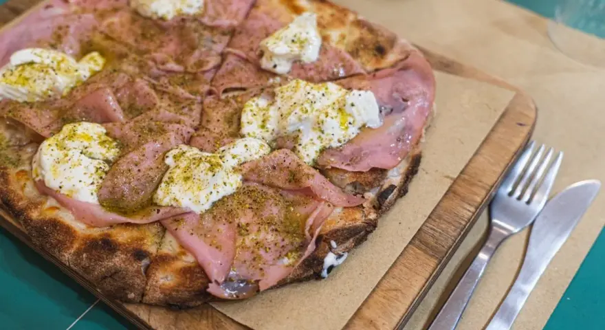 Pizza con mortadela y burrata cremosa, espolvoreada con hierbas verdes y servida en tabla de madera.