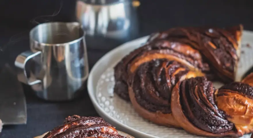 Deliciosa trenza de chocolate recién horneada, humeante junto a una taza de café.