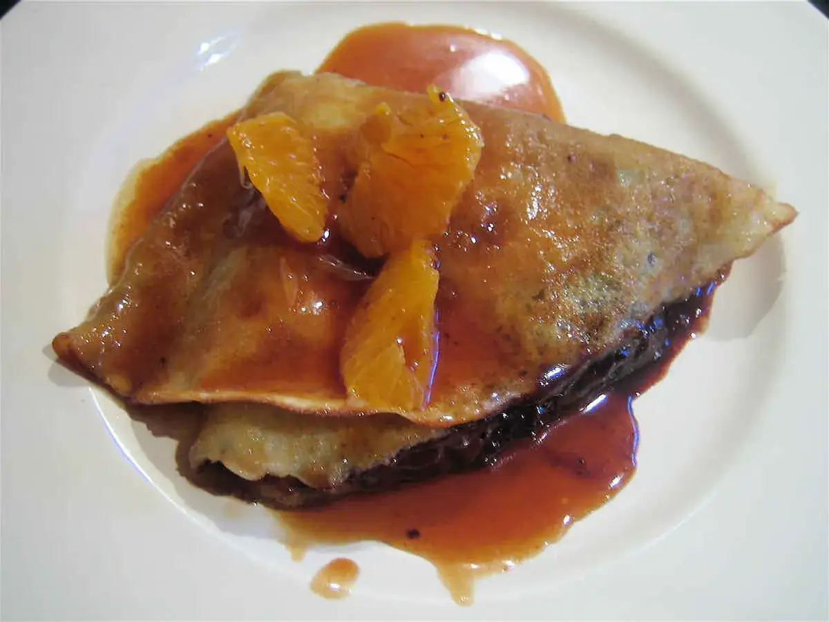 Crepe suzette doblada, cubierta con salsa de naranja y gajos de mandarina, servida en un plato blanco.
