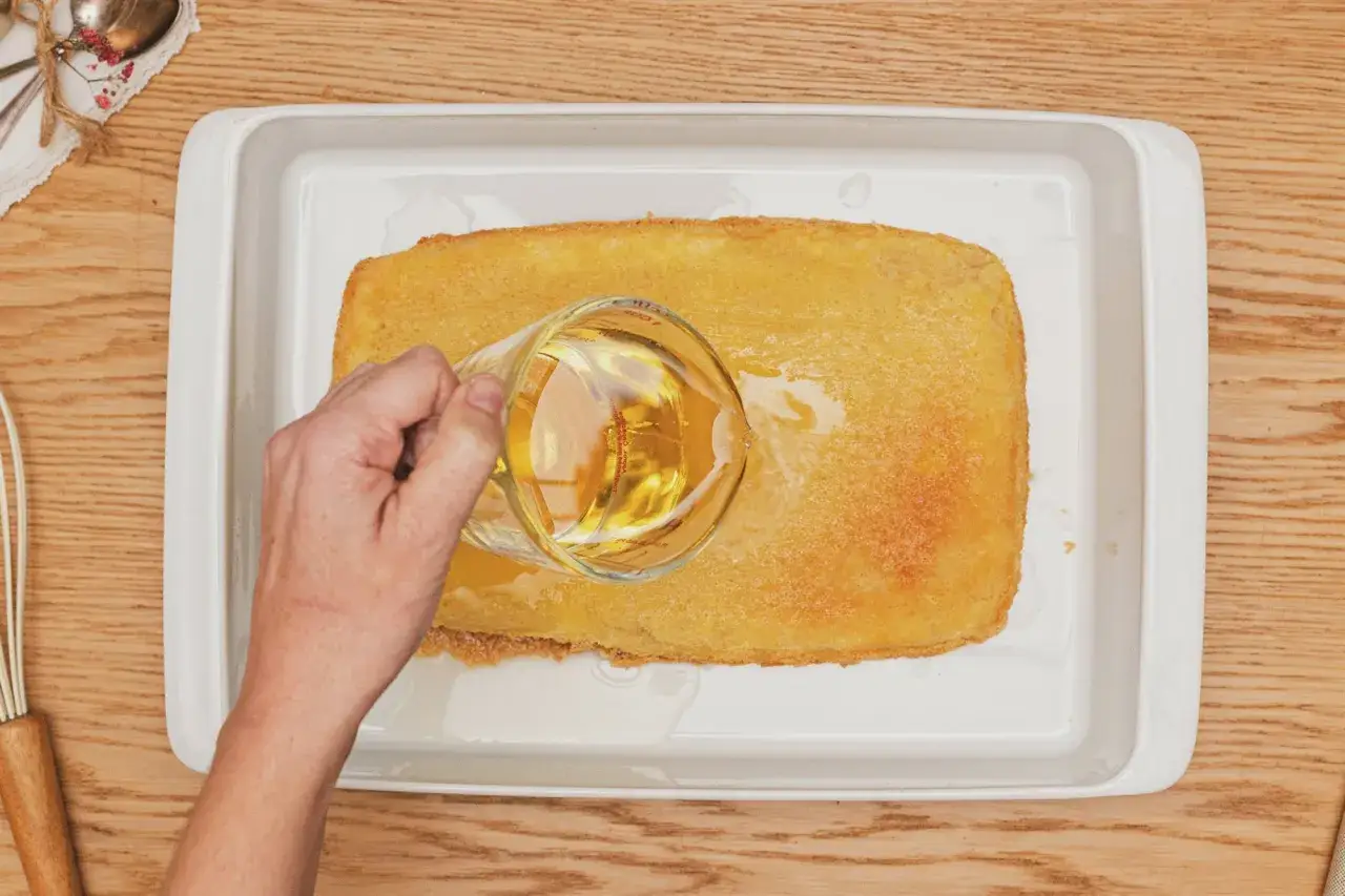 Mano vierte l&iacute;quido dorado sobre bizcocho borracho en bandeja blanca.