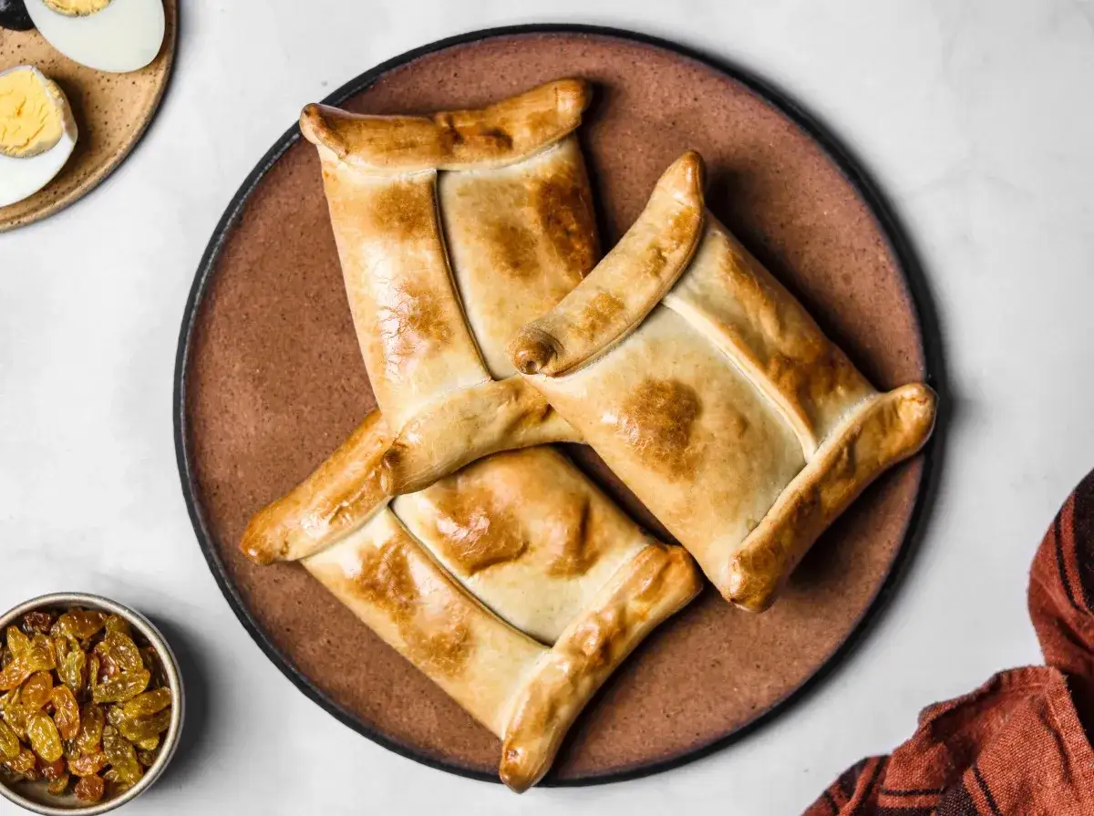 Tres empanadas chilenas doradas y crujientes dispuestas en un plato de barro. A un lado, un cuenco con pasas y un plato con huevo duro.