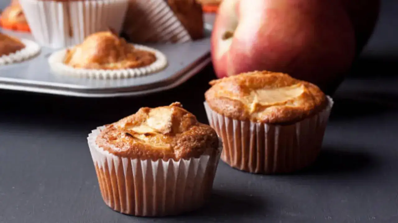 Dos deliciosos muffins de manzana reci&eacute;n horneados, con una manzana roja al fondo.