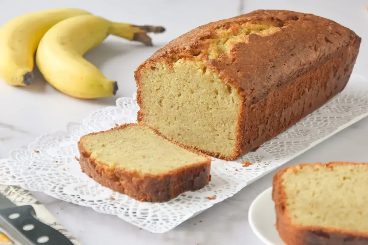 Un delicioso bizcocho de pl&aacute;tano reci&eacute;n horneado, con una rebanada cortada y dos pl&aacute;tanos amarillos al fondo.