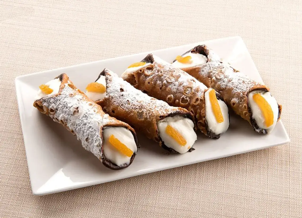 Cuatro cannoli siciliano, crujientes y cubiertos de az&uacute;car glas, rellenos de crema y decorados con naranja confitada.