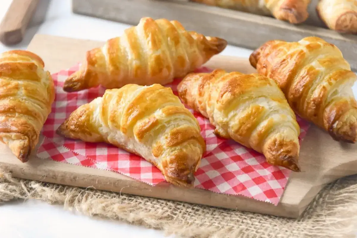 Seis croissants caseros dorados y hojaldrados reposan sobre una tabla de madera y un pa&ntilde;o a cuadros rojos.