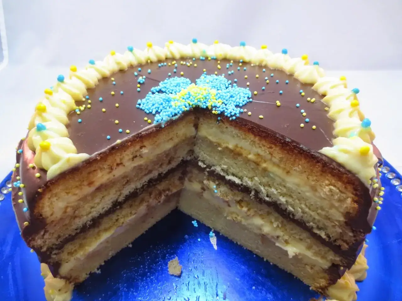 Pastel de capas con crema pastelera, cubierto de chocolate y decorado con chispas azules y amarillas.