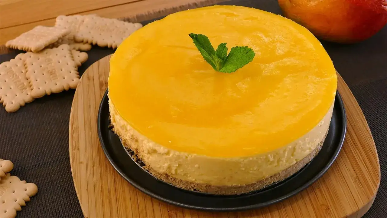 Deliciosa tarta de queso y mango, con una cobertura brillante y decorada con menta. Galletas tostadas al lado.