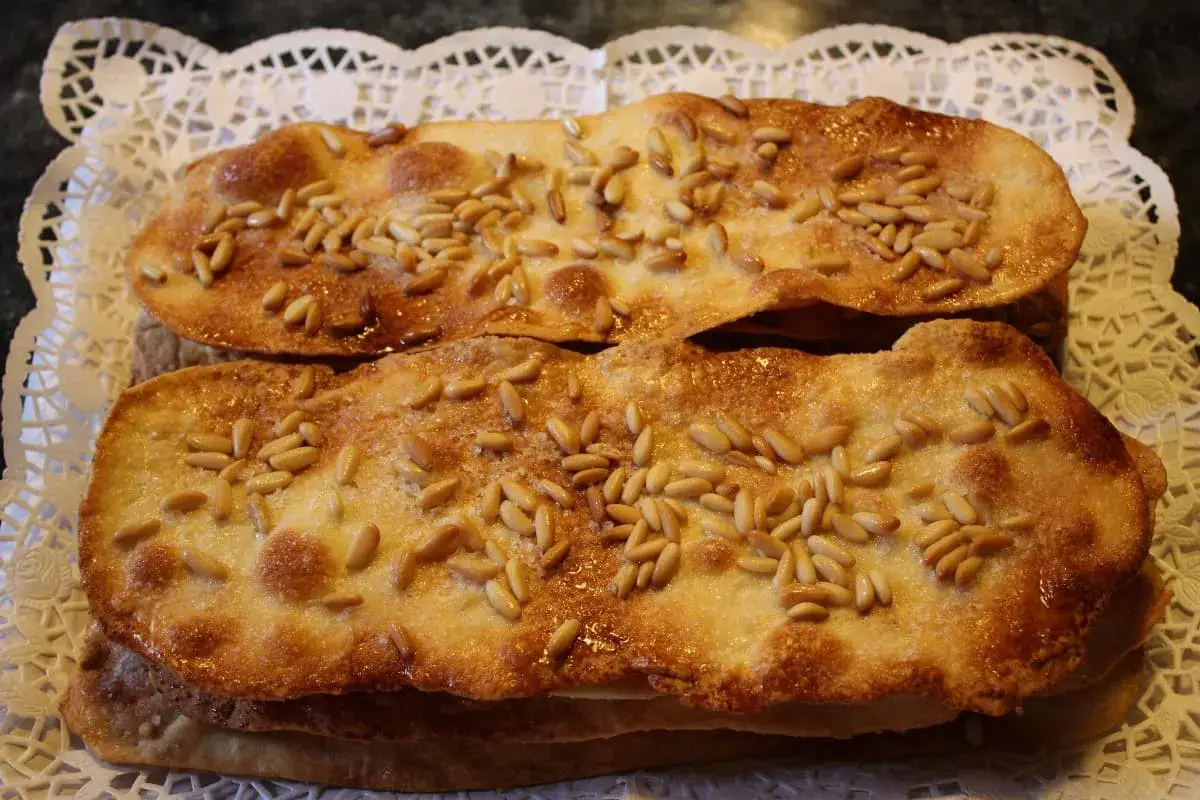 Dos coca de vidre doradas y crujientes, cubiertas de pi&ntilde;ones y az&uacute;car, reposan sobre un encaje blanco.