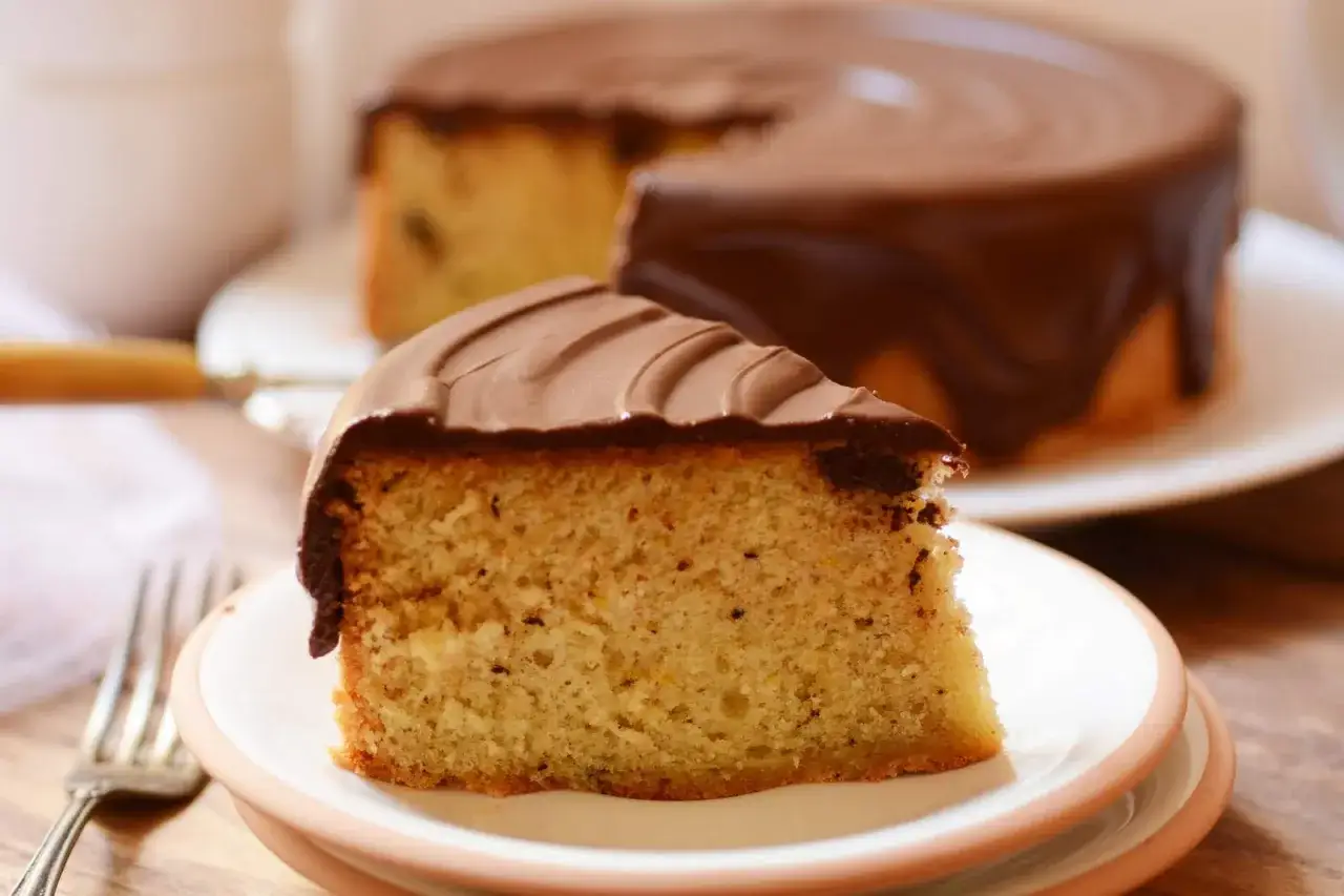 Un trozo de bizcocho esponjoso con una deliciosa cobertura de chocolate para bizcocho, con el bizcocho entero al fondo.