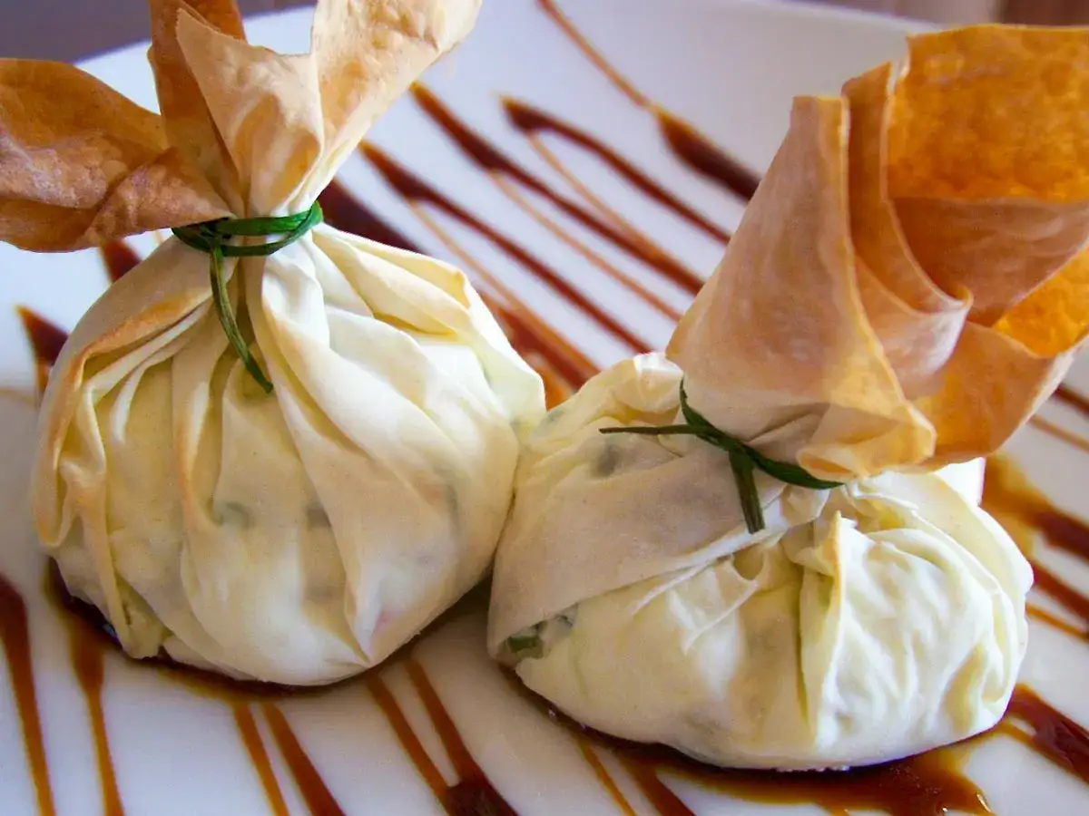Deliciosos saquitos de pasta filo rellenos de queso, atados con hilo verde y servidos sobre un plato con salsa.