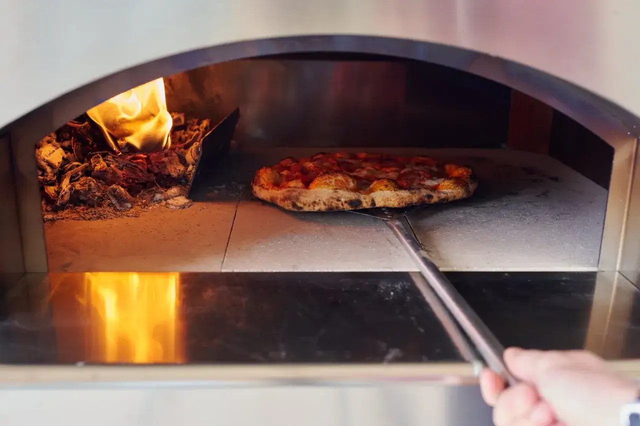 Una pizza masa madre dorada, lista para ser retirada del horno de le&ntilde;a, con brasas ardientes a un lado.