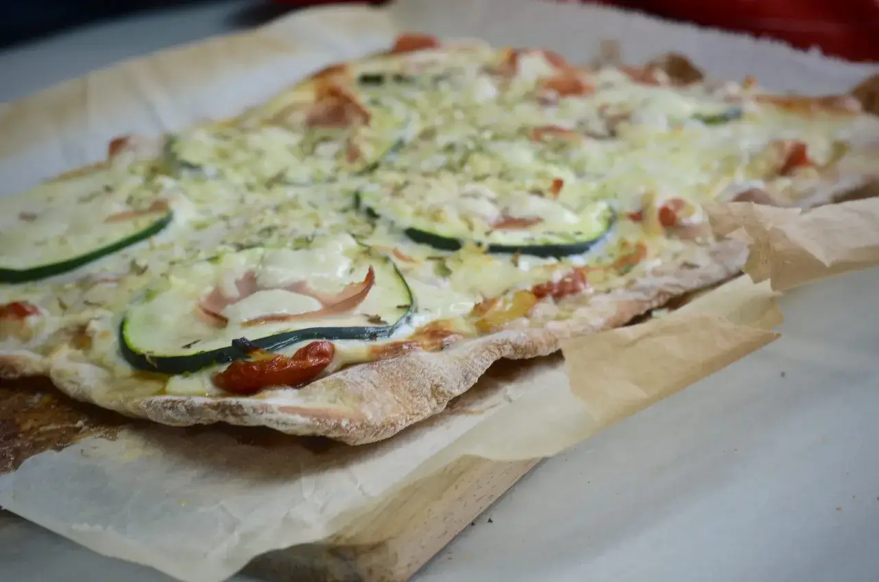 Pan pizza recién horneada con calabacín, tomate y jamón, sobre papel de horno.