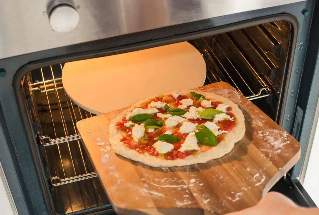 Pizza lista para hornear. Se ve la piedra para pizza dentro del horno, ya precalentado, y la pizza cruda en una pala de madera.