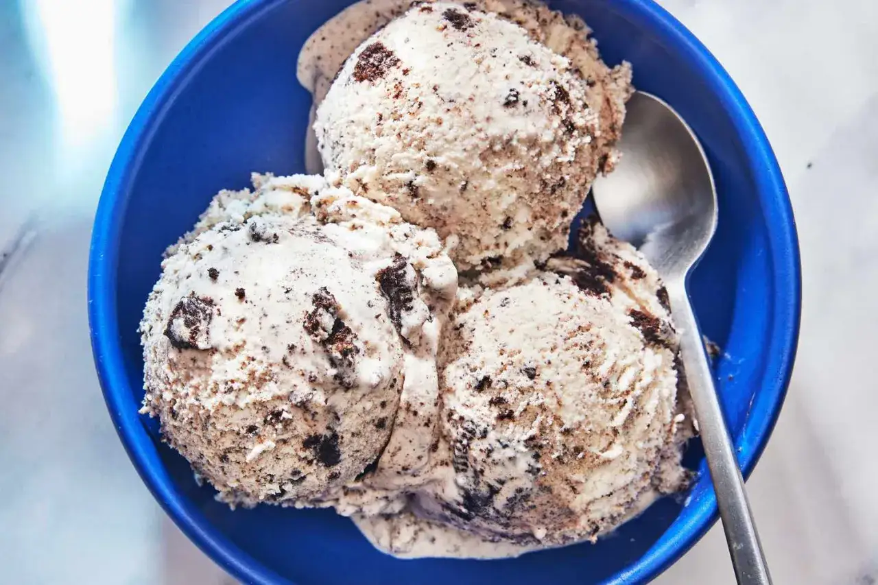 Un taz&oacute;n azul lleno de cremoso helado de galleta con trozos oscuros. Una cuchara plateada descansa a un lado, lista para disfrutar.