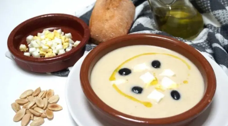 Un cuenco de mazamorra cordobesa con aceitunas, huevo duro y un chorrito de aceite. Acompaña un panecillo y almendras.